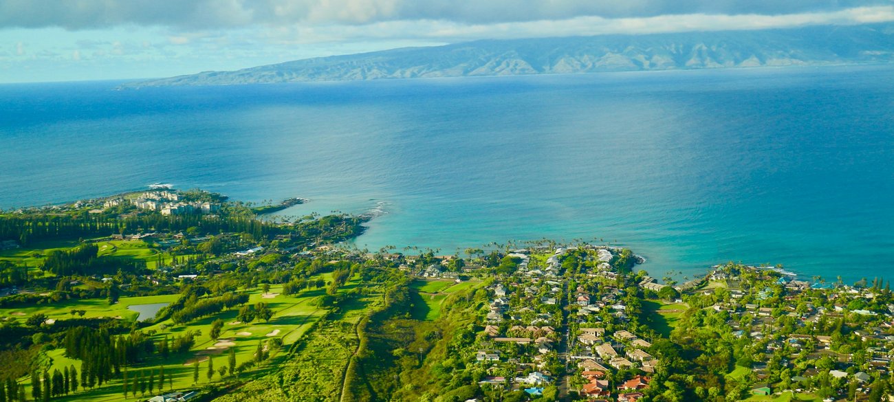 Aerial view of Napili Bay, Maui, highlighting the serene coastal town of Napili with its rich greenery and clear blue waters against a backdrop of mountains.