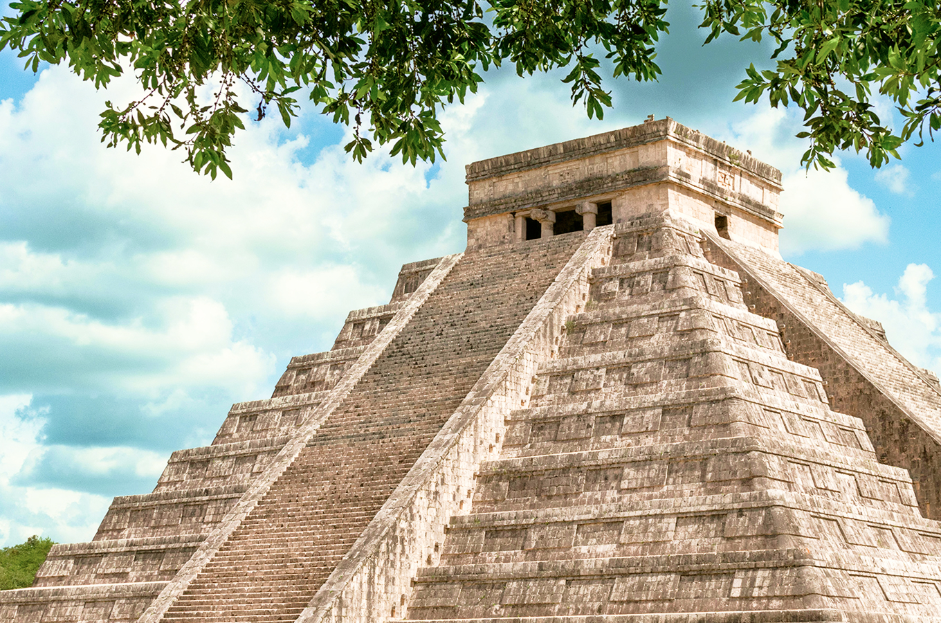 El Castillo pyramid at the Chichén Itzá Mayan ruins framed by lush foliage under a bright blue sky