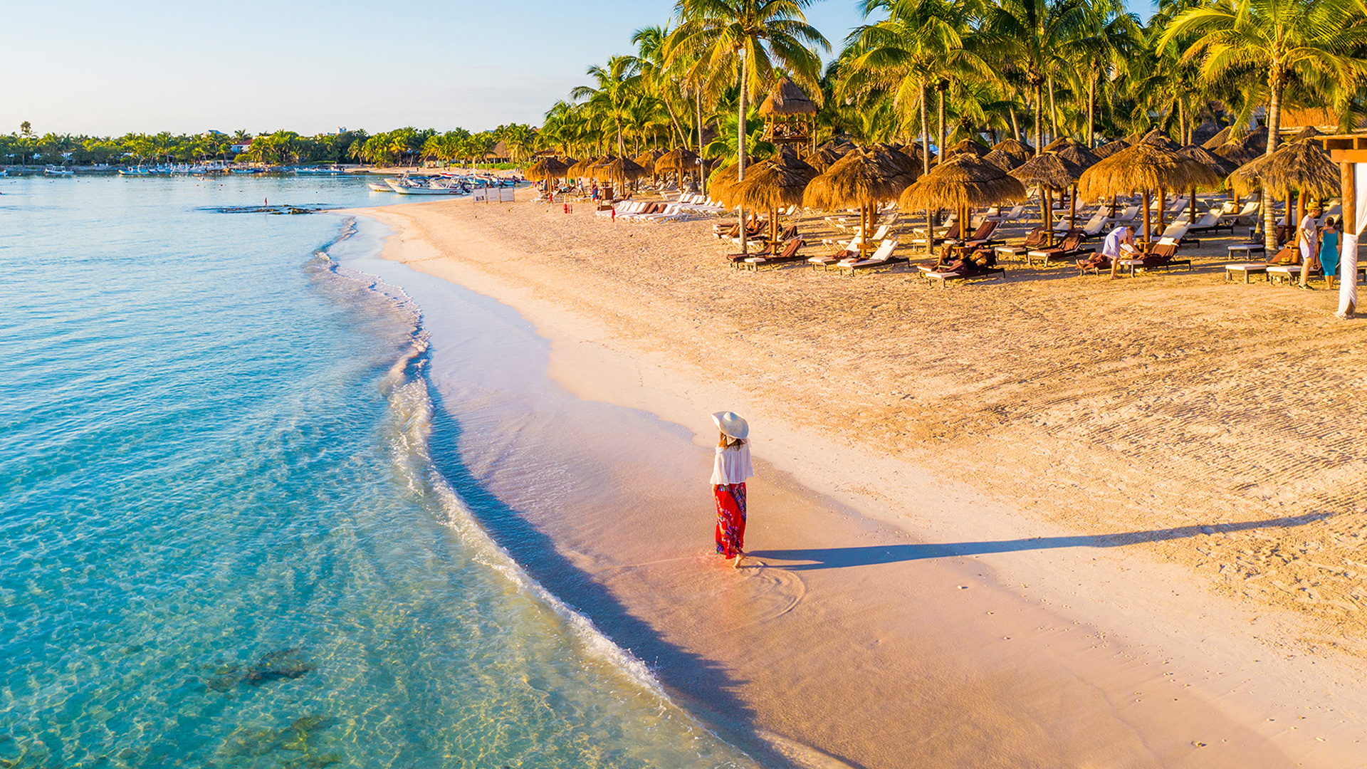 Woman in a sun hat strolling along a white-sand beach at golden hour, with palm-thatched palapas and lounge chairs lining the shore in Cancún
