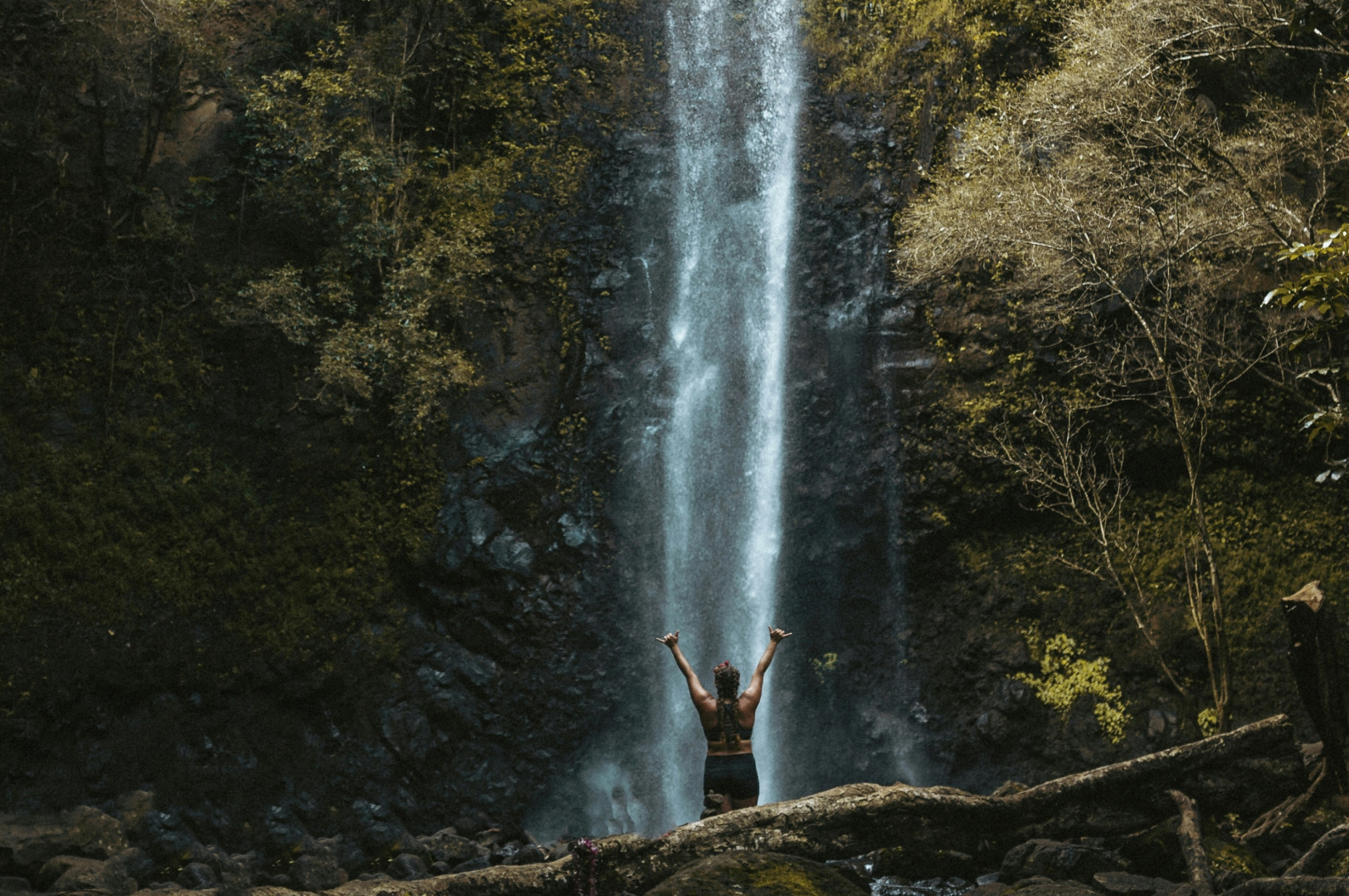 A person stands with arms raised in front of a towering waterfall surrounded by lush vegetation in Kauai, Hawaii.