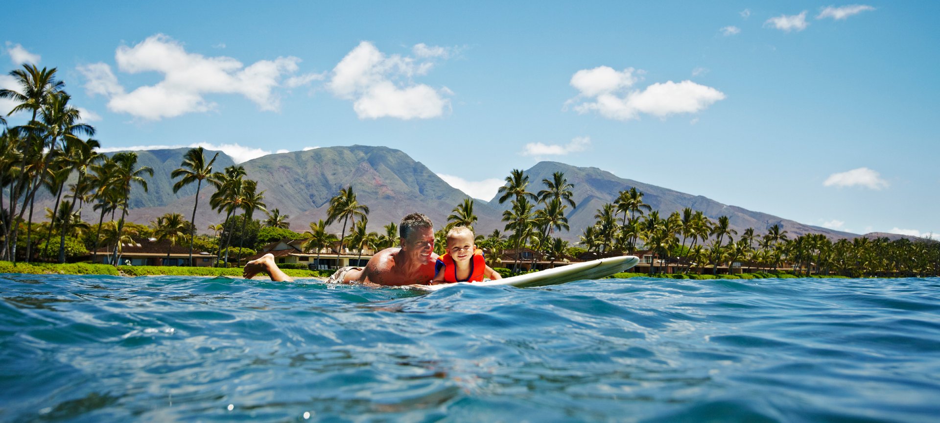 A father and his son on a surfboard paddle through the waters of Maui, Hawaii, with tropical palms and the mountains as backdrop.