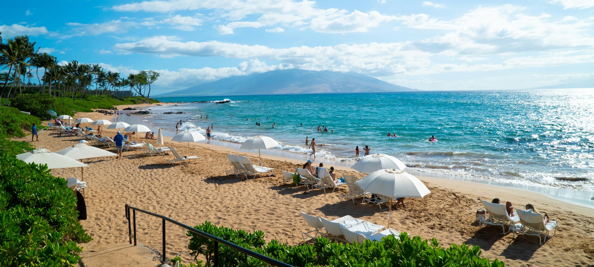 Beachgoers relax under white umbrellas on a sunny Maui shore with distant mountains.ont and a view of a hazy mountain in the distance.