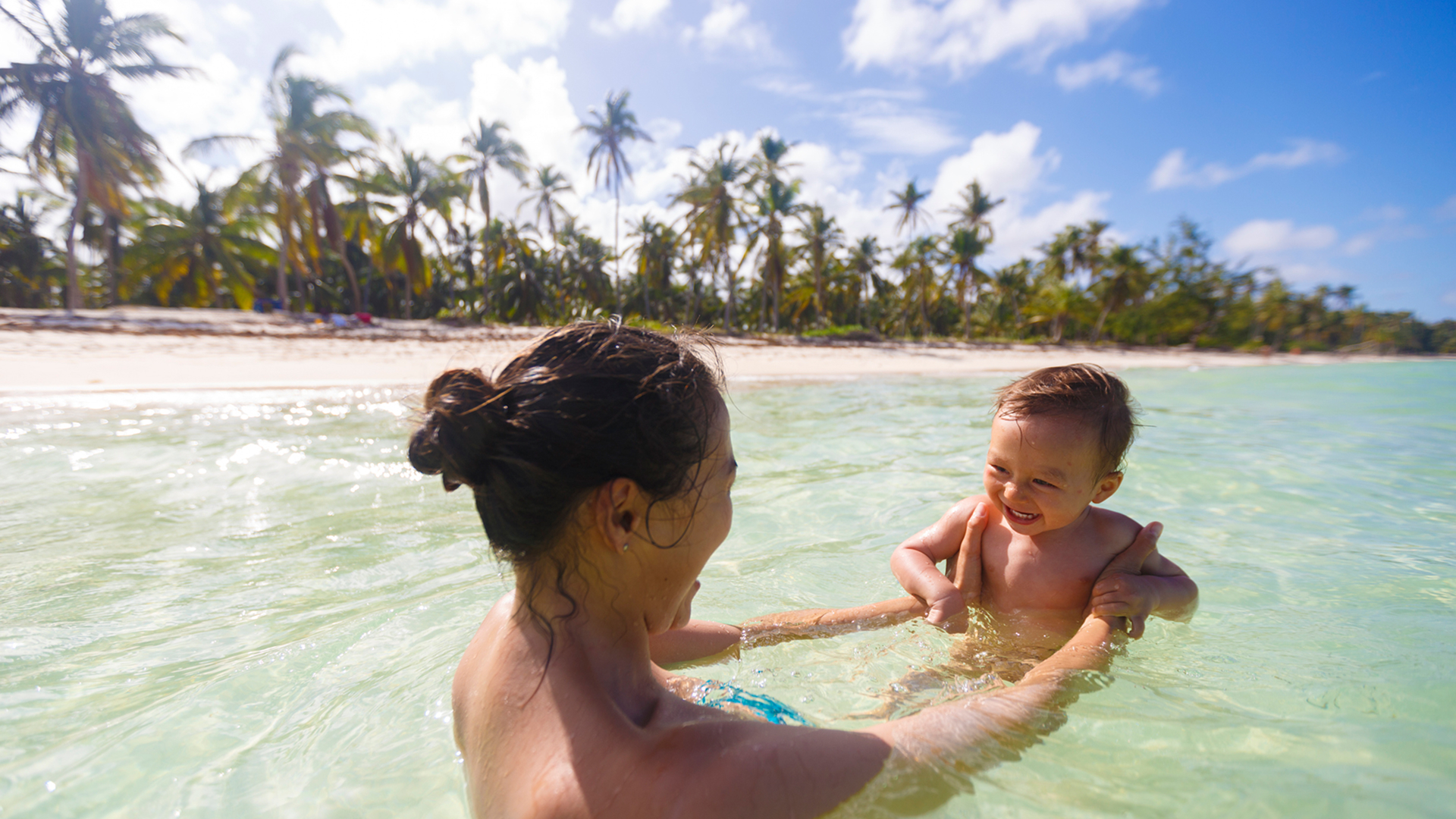 Mother lifting her toddler playfully in shallow, clear Caribbean waters along a palm-lined Punta Cana beach under a sunny sky