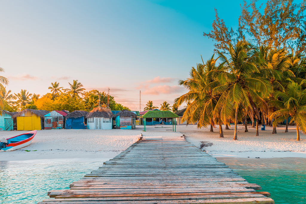 Wooden pier stretching into shallow turquoise Caribbean waters with paddle boats, thatched beach huts and palm trees lining the sandy shore in Punta Cana